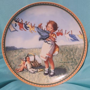 May include: A decorative plate featuring a young girl hanging laundry on a clothesline. The plate has a detailed illustration with a rural landscape background, a basket, and two dolls. The plate has a gold and green border.