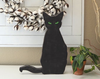 May include: A black cat silhouette with glowing green eyes sits on a white surface. Behind it is a white framed square with a cotton wreath. A potted plant is to the right.