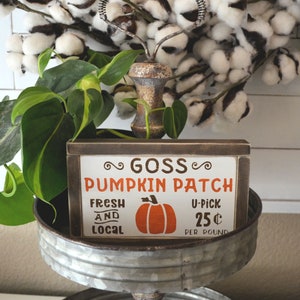 May include: A rustic wooden sign with the words "Goss Pumpkin Patch" in orange and brown. The sign also features a pumpkin graphic and the words "Fresh and Local". A metal whisk and cotton stems are in the background.