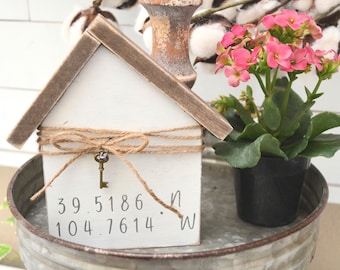 Puede incluir: Un letrero de madera blanco con forma de casa con un lazo de cuerda y un pequeño dije de llave. El letrero tiene las coordenadas 39.5186 N 104.7614 W escritas en él. El letrero está sentado en una bandeja de metal con una planta de flores rosadas en una maceta negra.