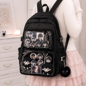 Puede incluir: Una mochila negra con dos bolsillos frontales transparentes llenos de varios pines circulares de personajes, pequeñas figuras de peluche y un pompón negro esponjoso sujeto a un lado. La bolsa tiene correas ajustables y hebillas laterales.