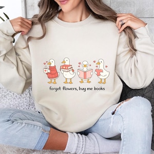 May include: Cream-coloured sweatshirt with a cartoon design of four ducks, each with a different item: a cup, books, a book, and a heart. The text "forget flowers, buy me books" is printed below. Small hearts surround the ducks.