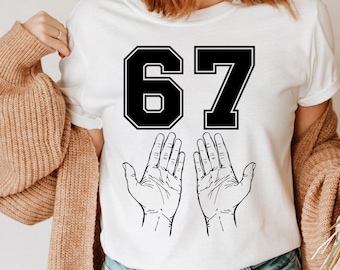May include: White t-shirt featuring the number "67" in a black, block-style font. Below the number are two open hands with detailed line drawings. The shirt is a casual, everyday garment.