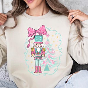 May include: A cream-colored sweatshirt featuring a whimsical Christmas design. The graphic includes a pink bow, a nutcracker soldier in pink and teal, and a decorated Christmas tree with pink ornaments, all within a teal outline.