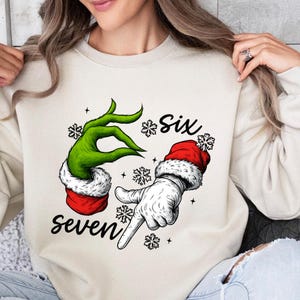 Può includere: Felpa color crema con una mano verde dal design Grinch e una mano guantata bianca con cappello da Babbo Natale. Le parole "Six" e "seven" sono scritte in corsivo nero, con fiocchi di neve e stelle.