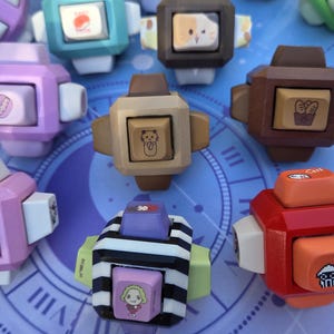 Könnte beinhalten: Eine Sammlung farbenfroher, würfelförmiger Tastenkappen mit verschiedenen Designs und Illustrationen. Die Tastenkappen sind in den Farbtönen Rosa, Braun, Grün und Rot gehalten, mit weißen und beigen Akzenten. Einige zeigen Cartoon-Figuren und Essenssymbole.