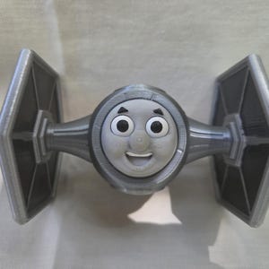 Può includere: Un giocattolo grigio stampato in 3D che combina il design di un caccia TIE di Star Wars con la faccia di Thomas il Trenino. Il giocattolo ha una faccia sorridente e due ali angolate su entrambi i lati.