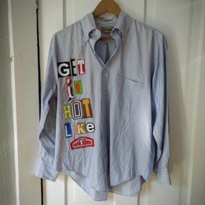 May include: Light blue striped button-down shirt with red accent stripes. The shirt features the text "GET IT HOT LIKE" in colorful, patchwork-style lettering, with a Papa John's logo. The shirt is hanging on a wooden hanger.
