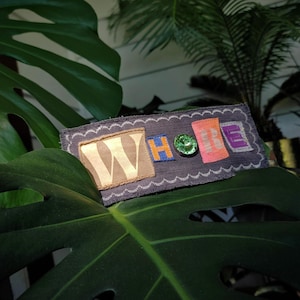May include: A rectangular patch with the word "WHOLE" spelled out in colorful, textured letters. The patch is dark blue with a white wavy border and a green gem. The patch is resting on a large green leaf.