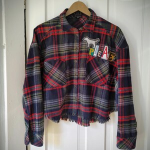 May include: A cropped, long-sleeved plaid shirt in red, navy, and green. The shirt features a frayed hem and two front pockets. Patches on the chest spell "PLEASE" and feature a dog silhouette. The shirt is hanging on a wooden hanger.