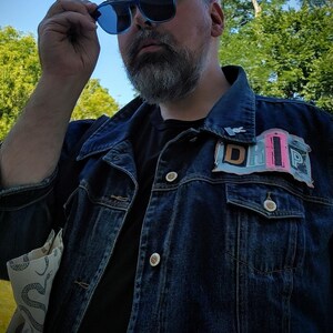 Puede incluir: Chaqueta vaquera azul oscuro con un parche "DRIP" en rosa, azul y blanco. La chaqueta tiene botones plateados y se lleva sobre una camisa negra. La persona lleva gafas de sol azules.