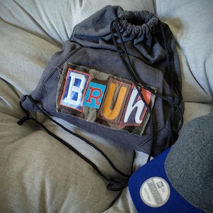 May include: A gray drawstring backpack with the word "BRUH" appliqued on the front in colorful, block letter patches. The backpack has black drawstrings and is paired with a blue and gray baseball cap.
