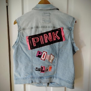 May include: Light blue denim vest with the words "PINK PONY CLUB" appliqued on the back in various colors. The vest is sleeveless and has a collared neckline. The vest is hanging on a wooden hanger.