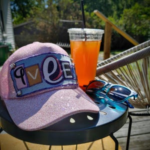 May include: A pink glitter baseball cap with the word "LOVE" in a patch, a clear plastic cup with an orange drink, and blue sunglasses on a small black table. The cap has a mesh back.