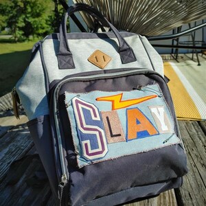 May include: A gray and navy blue backpack with a front pocket featuring the word "SLAY" in colorful letters and a lightning bolt. The backpack has a top handle, side pocket, and a brown diamond-shaped patch.