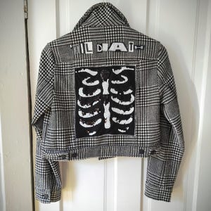 May include: Black and white houndstooth jacket featuring a sequined rib cage design on the back. The word "TILDA" is across the top. The jacket has a button closure and a collar.