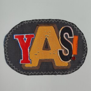 May include: An oval-shaped patch with the word "YAS!" in bold, colorful letters. The letters are in red, yellow, black, and orange, with a dark gray background and a white stitched border. The patch is likely for clothing or accessories.