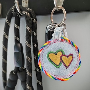 May include: A denim keychain with a rainbow-colored trim and two embroidered gold hearts. The keychain is attached to a set of keys and hangs next to a black and white dog leash.