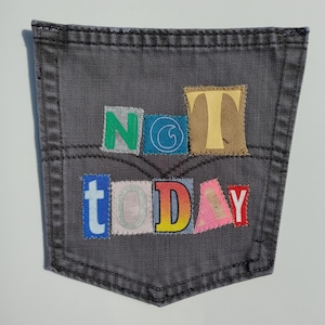 May include: A gray denim pocket with the words "NOT TODAY" appliquéd in colorful, patchwork-style letters. The pocket has a classic design with topstitching and a curved bottom edge, set against a white background.