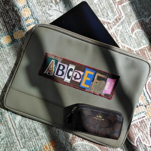 May include: A gray laptop sleeve with a colorful patch featuring the letters ABCDEF and a small brown Coach zip-around wallet. The laptop sleeve has a zipper closure and a laptop inside.