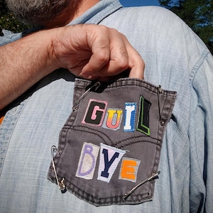 May include: A gray denim pocket with the words "GUIL BYE" appliqued in colorful letters. The pocket is secured to a light blue denim shirt with safety pins. The letters are in various colors, including pink, orange, and blue.