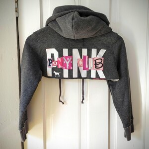 May include: A cropped gray hoodie with a hood and long sleeves. The word "PINK" is printed across the chest in white block letters, with the words "PONY CLUB" in colorful patches. A small white dog logo is visible.