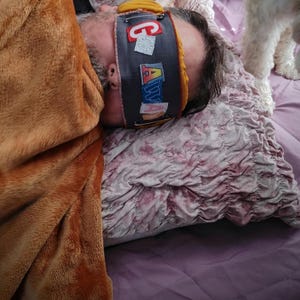 May include: A person wearing a grey sleep mask with the word "GALAXY" in colourful letters. The person is lying on a ruffled pink pillow and covered with a brown blanket. A white dog is in the background.