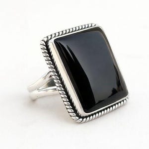 May include: A rectangular black onyx ring with a silver band. The ring features a polished black stone set within a rope-like border. The band splits into two at the base of the stone.