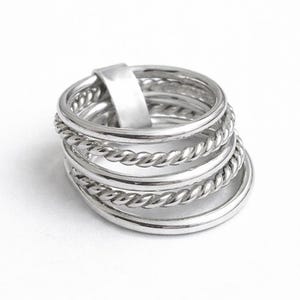 Peut inclure: Plusieurs bagues argentées de différents styles. Certaines présentent un motif torsadé, d'autres sont lisses. Les anneaux sont présentés sur un fond blanc, mettant en valeur leur éclat métallique.