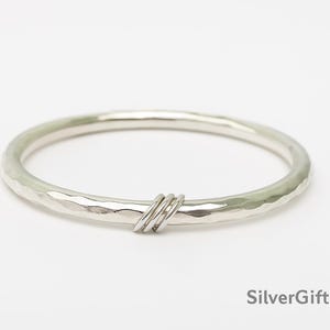 Può includere: Un bracciale a cerchio in argento con una texture martellata e un design semplice. Il bracciale presenta una fascia strutturata e un piccolo dettaglio decorativo di tre anelli d'argento. Il bracciale è mostrato su sfondo bianco. Il testo "SilverGiftLuxury" è visibile.