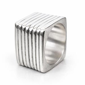Puede incluir: Un anillo plateado con un diseño cuadrado. El anillo presenta una serie de crestas verticales y paralelas en la superficie exterior, creando un efecto texturizado. El anillo está hecho de un material metálico brillante.