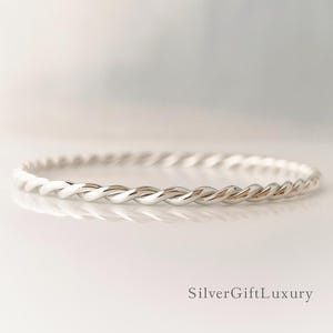 May include: A silver-colored bangle bracelet with a twisted rope design. The bracelet is circular and reflects light, creating highlights and shadows. The bracelet is displayed on a white surface. The text "SilverGiftLuxury" is in the bottom right corner.