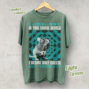 Op de afbeelding: Lichtgroen T-shirt met een rattenafbeelding en de tekst "IN THIS SINFUL WORLD" en "I DESIRE ONLY CHEESE". Het ontwerp heeft een turquoise en zwarte mandala-stijl achtergrond. Het shirt is een comfort kleur.