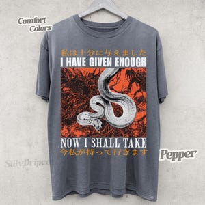Può includere: T-shirt grigio scuro con grafica di un serpente bianco su sfondo arancione. La maglietta presenta le scritte "I HAVE GIVEN ENOUGH" e "NOW I SHALL TAKE" in bianco, con testo giapponese sopra e sotto.