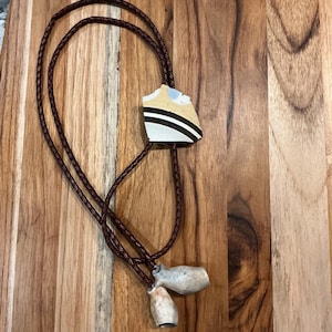 May include: A bolo tie with a brown braided cord and a rectangular pendant. The pendant features a tan, white, and black design. Two small, light-colored, cylindrical ornaments hang from the cord.