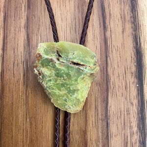 May include: A bolo tie featuring a rough-cut, light green stone pendant. The stone is suspended from a dark brown, braided leather cord. The background is a wooden surface with a natural grain pattern.