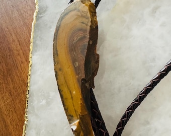 May include: A bolo tie featuring a polished, irregularly shaped stone pendant with brown, yellow, and tan hues. The pendant is suspended from a braided, dark brown leather cord, creating a rustic and natural aesthetic.