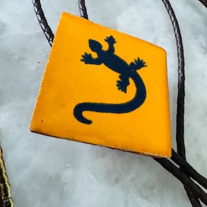 May include: A bolo tie featuring a square, orange pendant with a dark blue lizard silhouette. The pendant is attached to a braided brown leather cord. The lizard design is a striking contrast against the orange background.