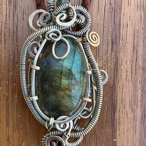 May include: A handcrafted bolo tie featuring a large, oval labradorite stone with iridescent green and blue hues, set in a silver wire wrap. The pendant is suspended from a braided brown leather cord, creating a unique accessory.