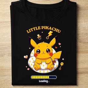 May include: Black t-shirt featuring a cartoon Pikachu character with the text "Little Pikachu". The design includes a pacifier, lightning bolt, and a loading bar graphic. The shirt is folded.
