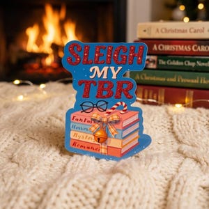 Może przedstawiać: Niebiesko-czerwony znak dekoracyjny z napisem "SLEIGH MY TBR" i stos książek. Książki są oznaczone jako "Fantasy, Horror, Mystery, Romance". Widoczne są również okulary i laska cukrowa.