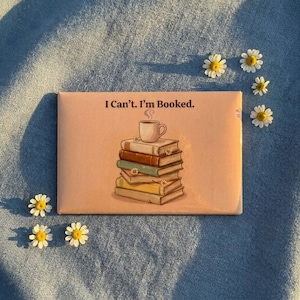 May include: A rectangular peach-colored magnet featuring the text I Can't. I'm Booked. above an illustration of a stack of five books with a steaming coffee mug on top. Small white daisy flowers are scattered around the stack of books.