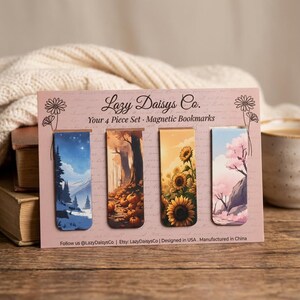 Puede incluir: Un juego de cuatro marcapáginas magnéticos en una tarjeta de presentación rosa con la etiqueta Lazy Daisys Co. Los marcapáginas muestran ilustraciones de un paisaje montañoso nevado, un campo de calabazas, girasoles y un cerezo en flor.