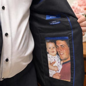 May include: Black blazer with a blue trim and a photo inside the inner pocket. The photo shows a man and a baby. The blazer has a white shirt underneath. The blazer has the text "AT102" and "SLIM FIT PERFORMANCE" on a tag.