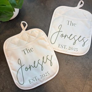 May include: Two off-white quilted oven mitts with a loop for hanging. Each mitt features the text "The Joneses EST. 2025" in a script font. The mitts are designed for kitchen use.