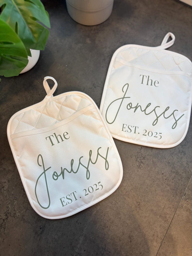 May include: Two off-white quilted oven mitts with a loop for hanging. Each mitt features the text "The Joneses EST. 2025" in a script font. The mitts are designed for kitchen use.