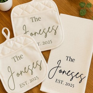 May include: A kitchen set featuring two cream-colored quilted oven mitts and a matching tea towel. Each item is printed with "The Joneses" and "EST. 2025" in a stylish script font, perfect for a housewarming or wedding gift.