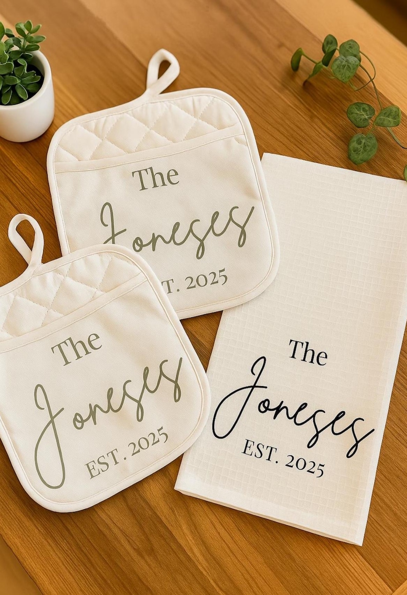 May include: A kitchen set featuring two cream-colored quilted oven mitts and a matching tea towel. Each item is printed with "The Joneses" and "EST. 2025" in a stylish script font, perfect for a housewarming or wedding gift.