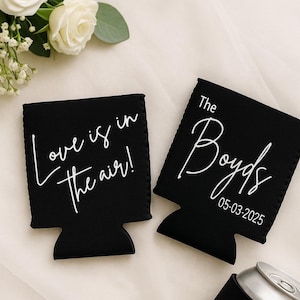 May include: Black drinks insulators with white text. One reads "Love is in the air!" and the other says "The Boyds 05-03-2025". White flowers and greenery are in the background. The insulators are designed to keep drinks cold.