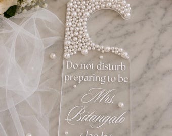 May include: A clear acrylic door hanger adorned with white pearls at the top. The text reads: Do not disturb preparing to be Mrs. Bitangalo 6/20/26. The hanger features a hook shape and is decorated with scattered individual pearls.